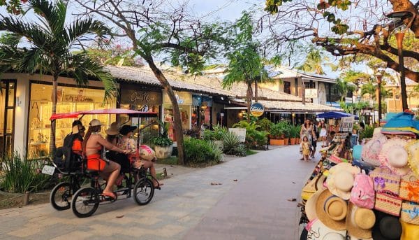 Vila da Praia do Forte com turistas passeando em bicicleta elétrica e diversas lojas de artesanato e moda ao longo do calçadão.