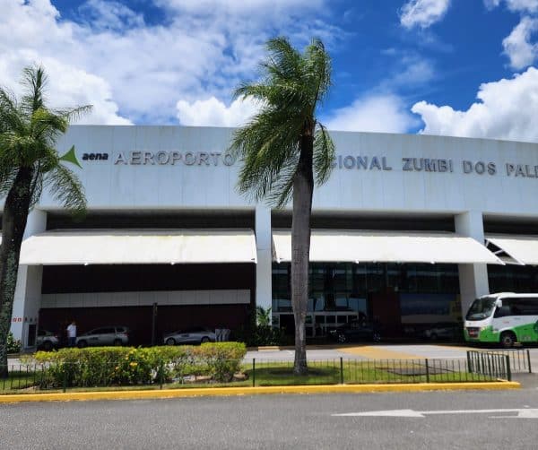 Fachada do Aeroporto Internacional Zumbi dos Palmares em Maceió, com palmeiras e céu azul ao fundo.