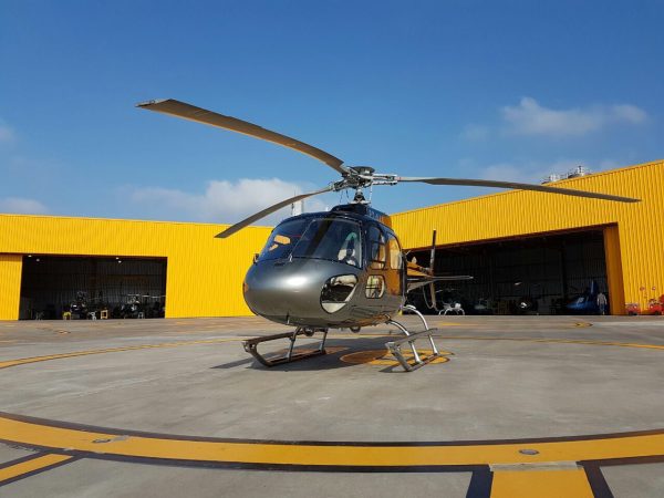 Helicóptero prateado estacionado em heliponto com hangar amarelo ao fundo, pronto para o embarque.