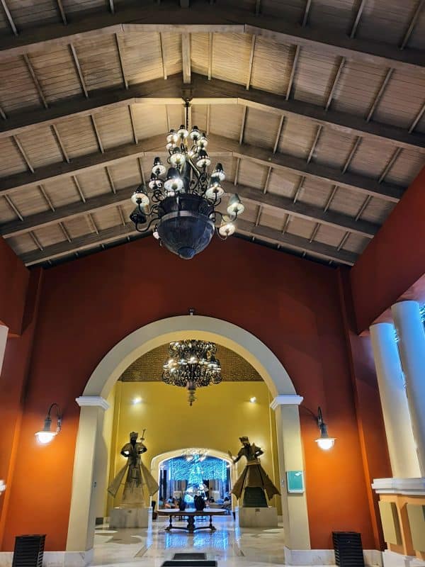 Hall de entrada do Iberostar Bahia com tetos altos de madeira, candelabros ornamentados e estátuas decorativas, oferecendo um ambiente sofisticado.