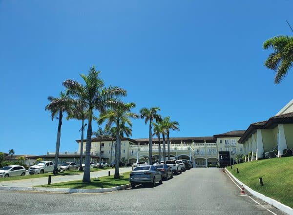 Entrada do Resort Costa do Sauípe com palmeiras e carros estacionados em um dia ensolarado.